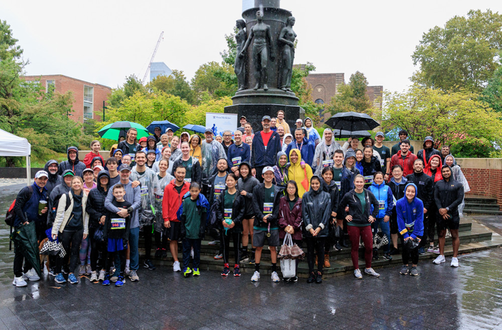 A group of race volunteers and runners poses on a rainy day at the base of a statue.